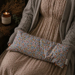Load image into Gallery viewer, Floral patterned lupin heat pillow on a person wearing a brown sweater with a candle and plant in the background.
