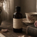Load image into Gallery viewer, A dark amber glass bottle of Chandler Perfumery 'Karsten &amp; Co.' Rose Garden essential oil with a black cap and cream label, styled on a rustic wooden table next to a vintage book, a floral teacup, and scattered rose petals.
