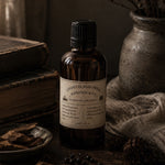 Load image into Gallery viewer, A dark amber glass perfume bottle with a black cap and a cream label reading 'Chandler Perfumery Karsten &amp; Co Cedarwood &amp; Bergamot', styled on a rustic wooden surface with books, a ceramic vase, and dried botanicals.
