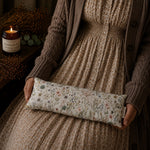 Load image into Gallery viewer, Person holding a floral-patterned lupin heat pillow with a candle and decorative elements in the background
