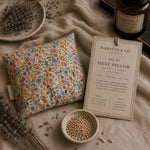Load image into Gallery viewer, Floral heat pillow with a label on a textured surface with lavender and grains.
