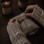 Load image into Gallery viewer, A person wearing a chunky cable-knit sweater wraps a folded linen cloth tied with twine in a cozy dark wood setting, with a lit candle and small plant in the background.
