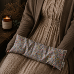 Load image into Gallery viewer, Person wearing a brown sweater holding a floral fabric lupin heat pillow next to a lit candle on a wooden surface.
