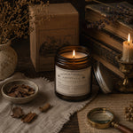 Load image into Gallery viewer, Pure essential oil candle in an amber jar with a label on a rustic setting with books and dried herbs - Handmade in Australia
