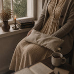 Load image into Gallery viewer, A person in a beige ribbed cardigan and textured dress sits by a window, holding a light beige lupin heat pillow. A mug, an open book, and a candle are on the table and windowsill.
