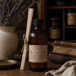Load image into Gallery viewer, Brown bottle with label and reed diffuser on a wooden surface with books and lavender in the background
