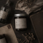 Load image into Gallery viewer, Organic Lavender Candle jar with label on a dark surface with bottle, paper, and flowers - hand poured in Australia by Karsten &amp; Co.
