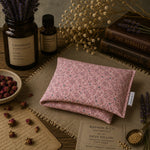 Load image into Gallery viewer, Pink floral heat pillow on a wooden surface with dried lavender, books, and herbs.
