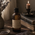 Load image into Gallery viewer, A bottle of Karsten &amp; Co Organic Lavender essential oil or perfume oil stands on a rustic wooden table, surrounded by dried lavender sprigs, an antique book, a lit candle in a metal holder, and a ceramic vase with dried flowers.
