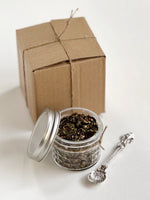 Load image into Gallery viewer, Open jar of tea leaves with a metal spoon and a cardboard box on a white background
